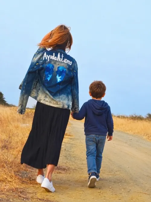 Paola Carlton walking hand in hand with a little boy down a dirt path, wearing her AyudaAutismo jacket